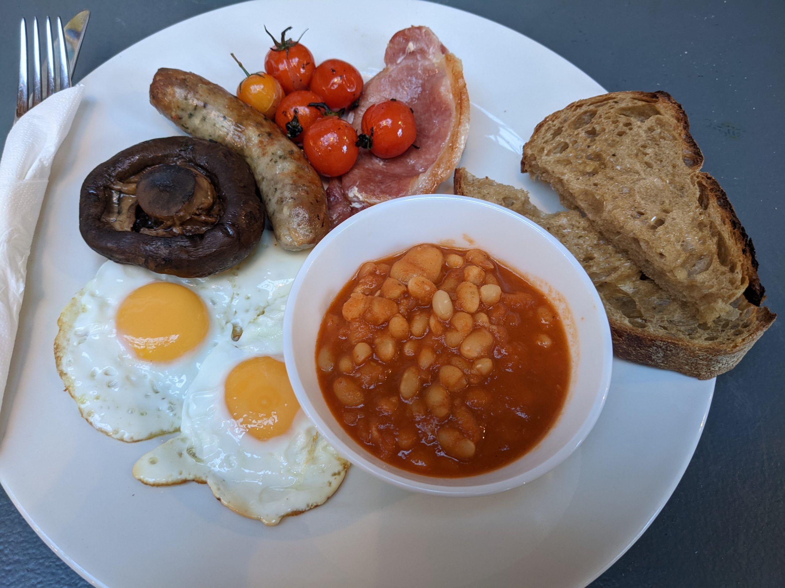 Na tanieri sú dve vajíčka, hríb, klobása, paradajky, slanina, fazuľová omáčka a opečený chlieb. Je to jedlo English Breakfast.