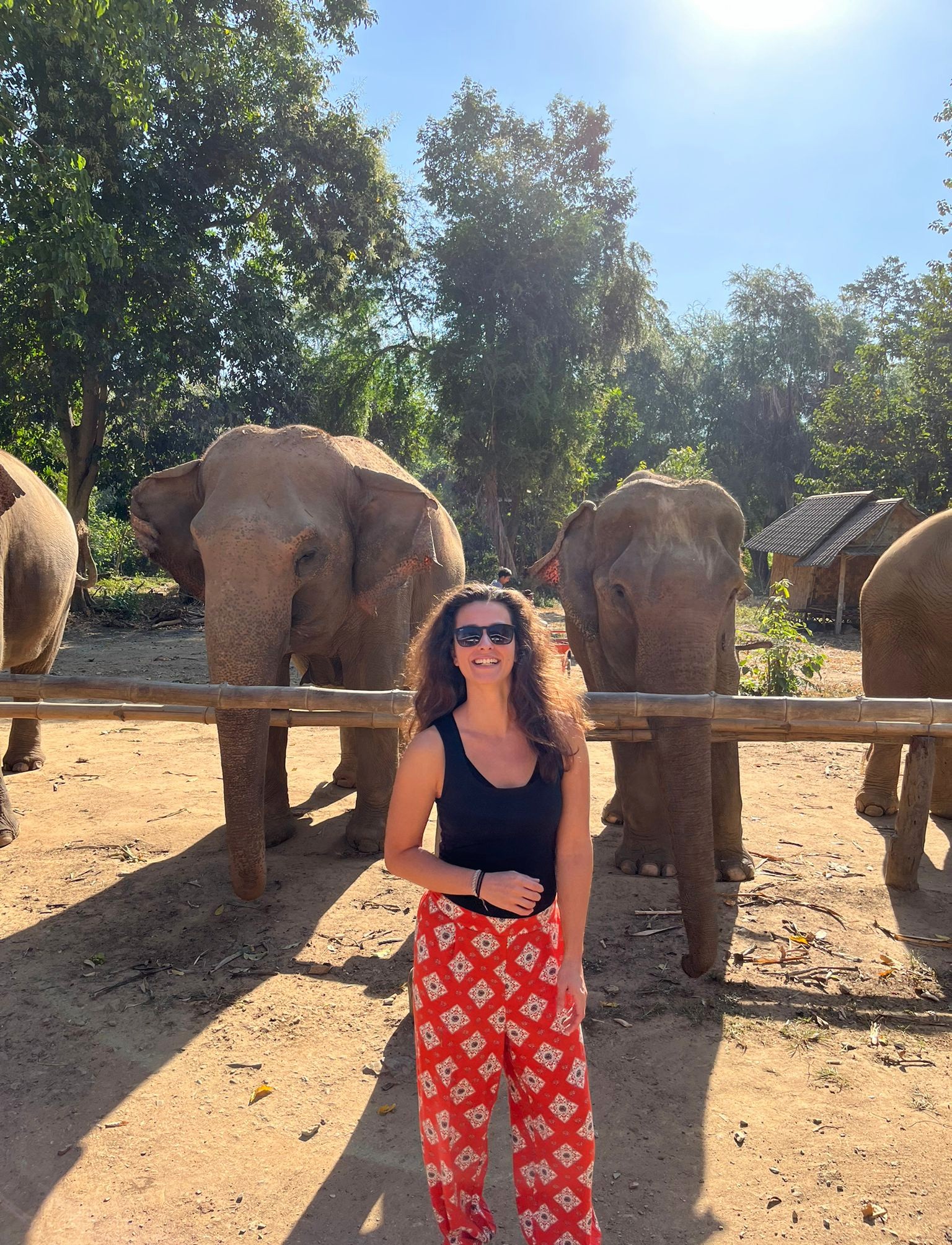 Žena v čiernom tričku a oranžovými nohavicami sa usmieva. Za ňou stoja slony.