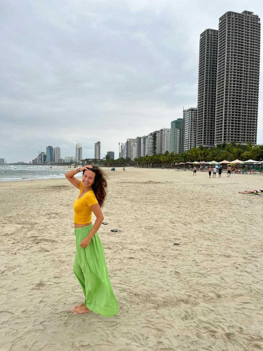 Dievča v zelenej sukni a oranžovom tričku stojí uprostred pláže a pózuje. Za ňou sú vysoké mrakodrapy.