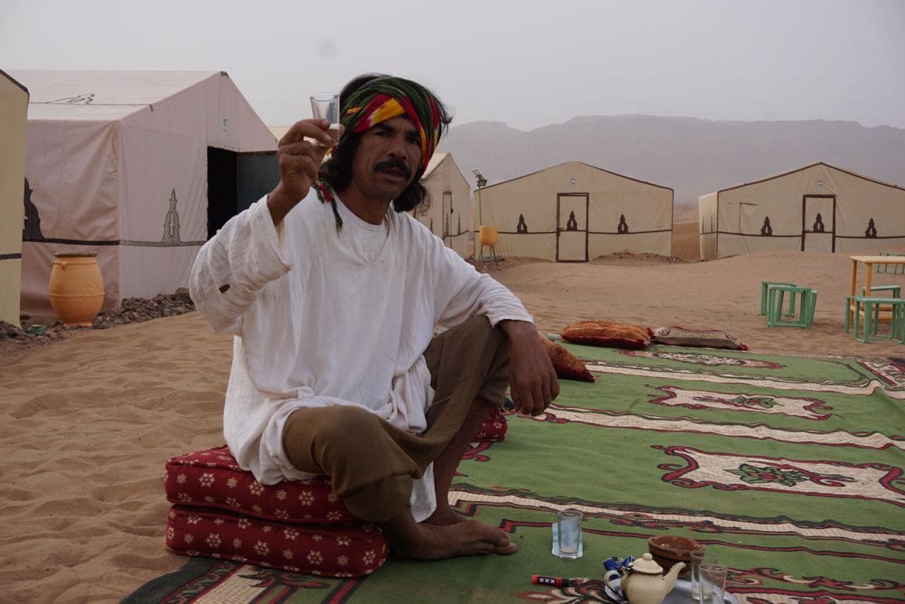 Chalp sediaci na koberčeku uprostred púšte. V ruke drží berberskú whiskey a v druhej brko s marockým hašišom.