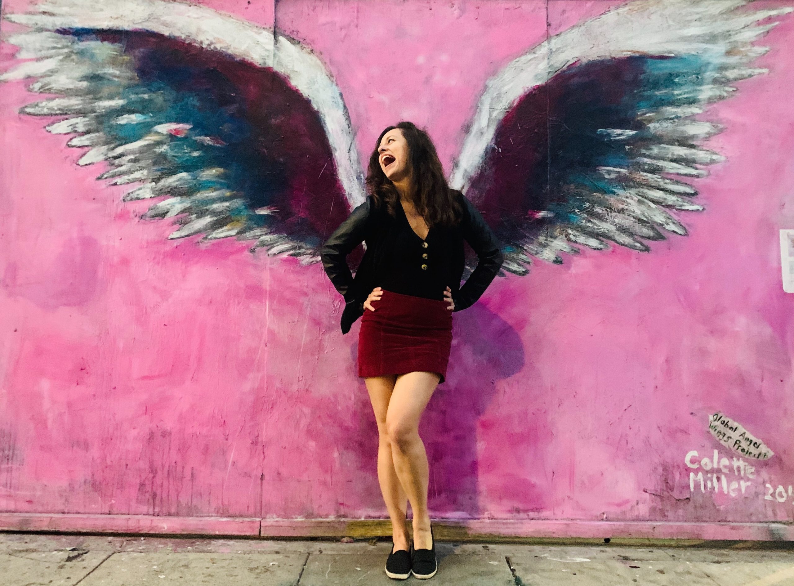 Dievča pózujúce s anjelskými krídlami pred rúžovou stenou.