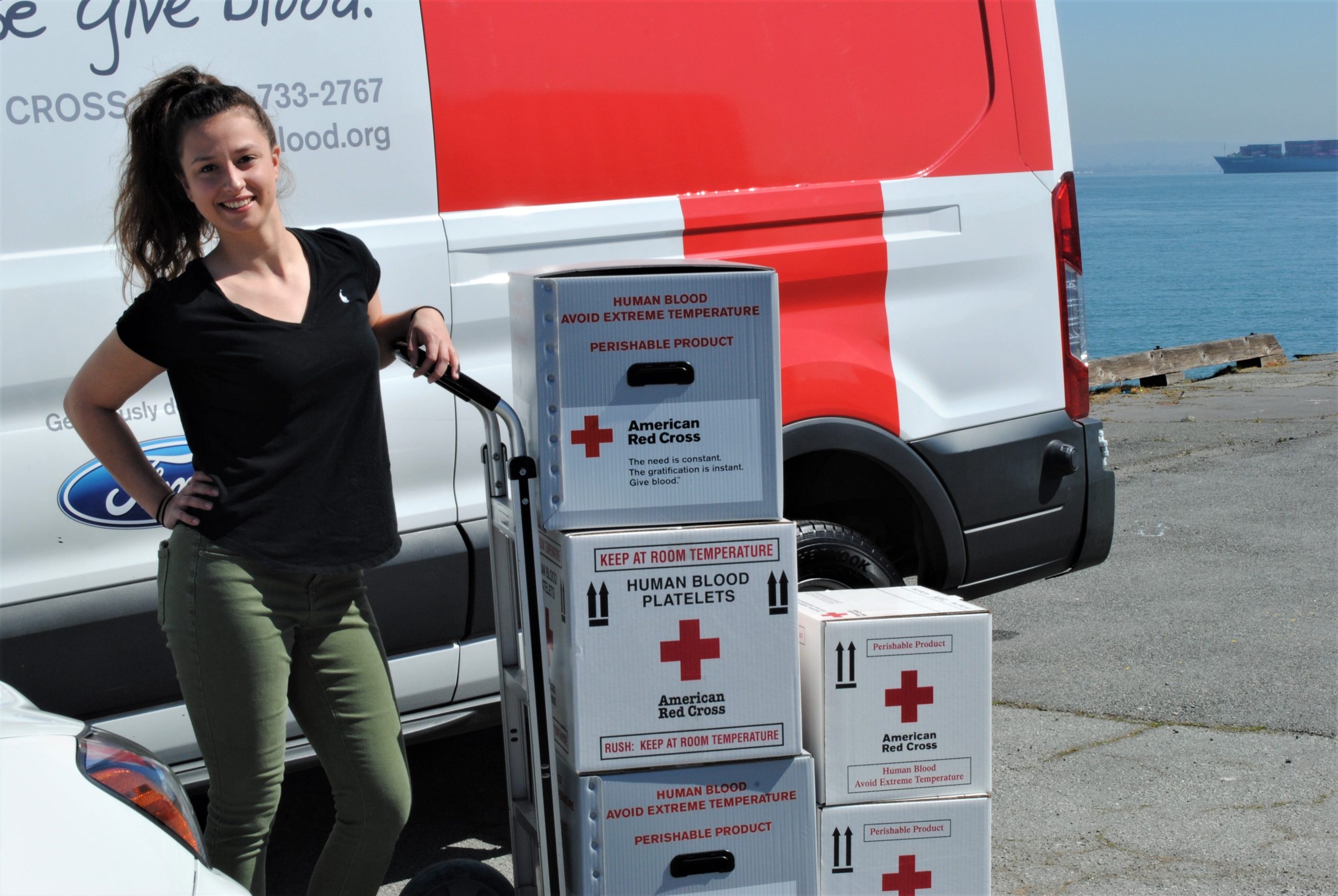 Dievča stojí okolo dvoch áut Červeného kríža a opiera sa o vozík s krabicami.