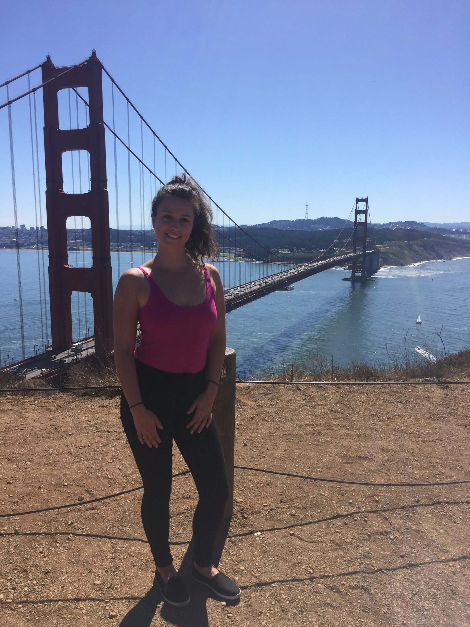 Dievča v rúžovom tričku a čiernych nohaviciach stojí pred mostom Golden Gate Bridge.