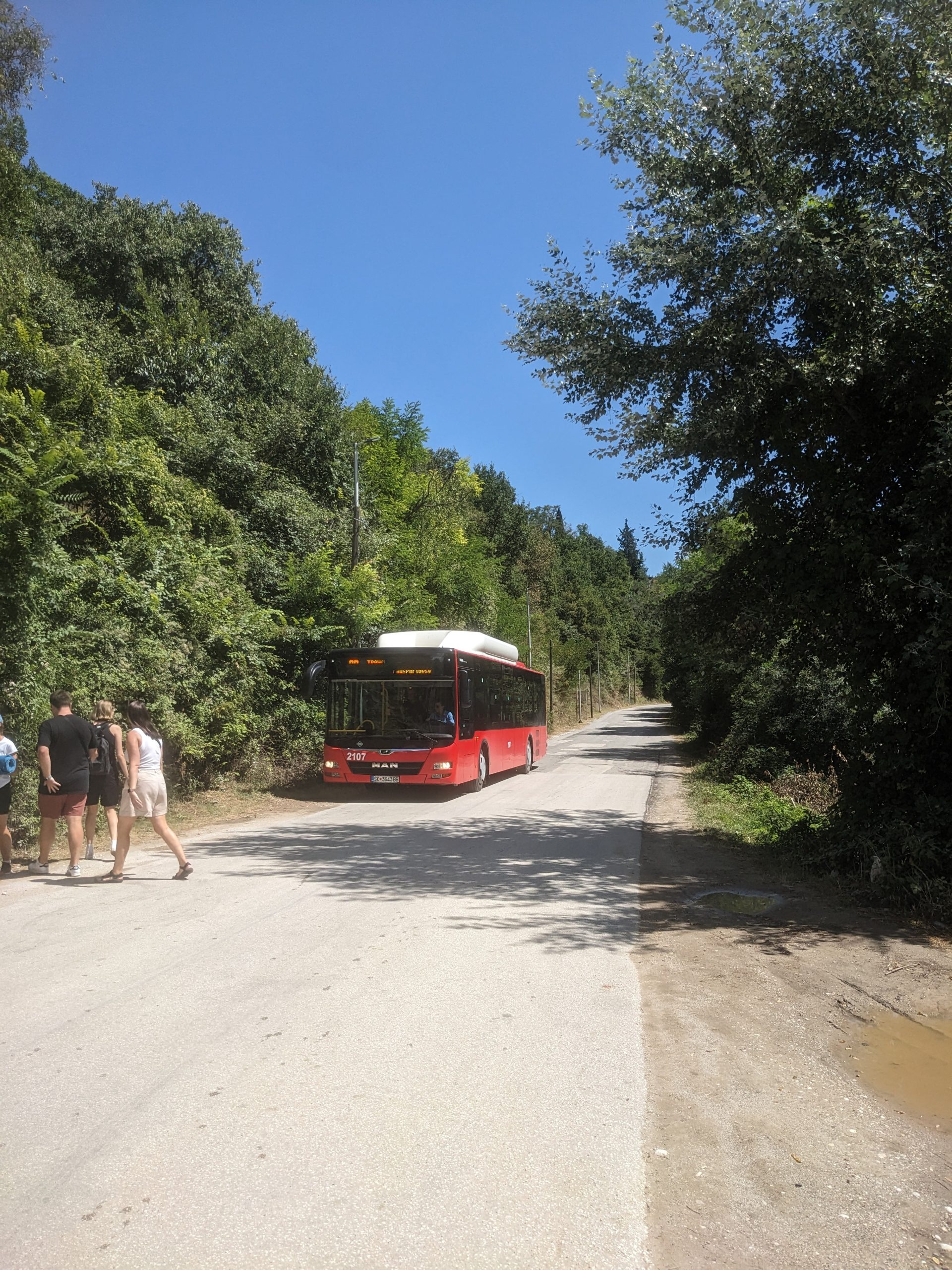 Červený autobus pri lese stojí na ceste. Pred ním kráčajú ľudia.