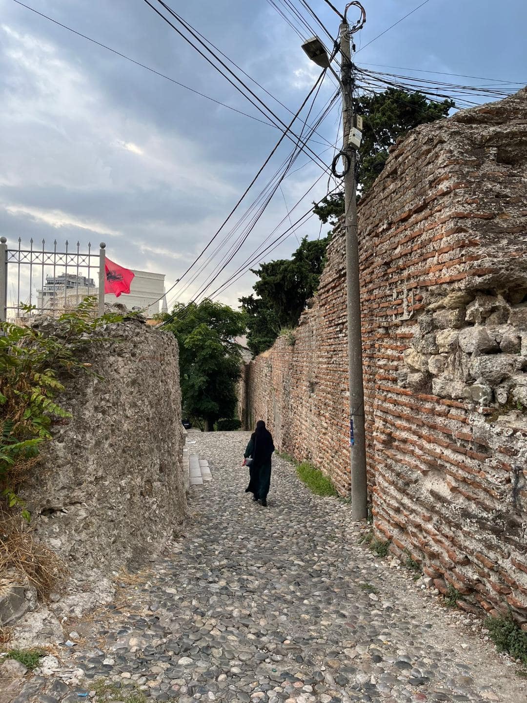 Moslimská žena prechádza okolo hradieb mesta. V pozadí veje albánska vlajka.