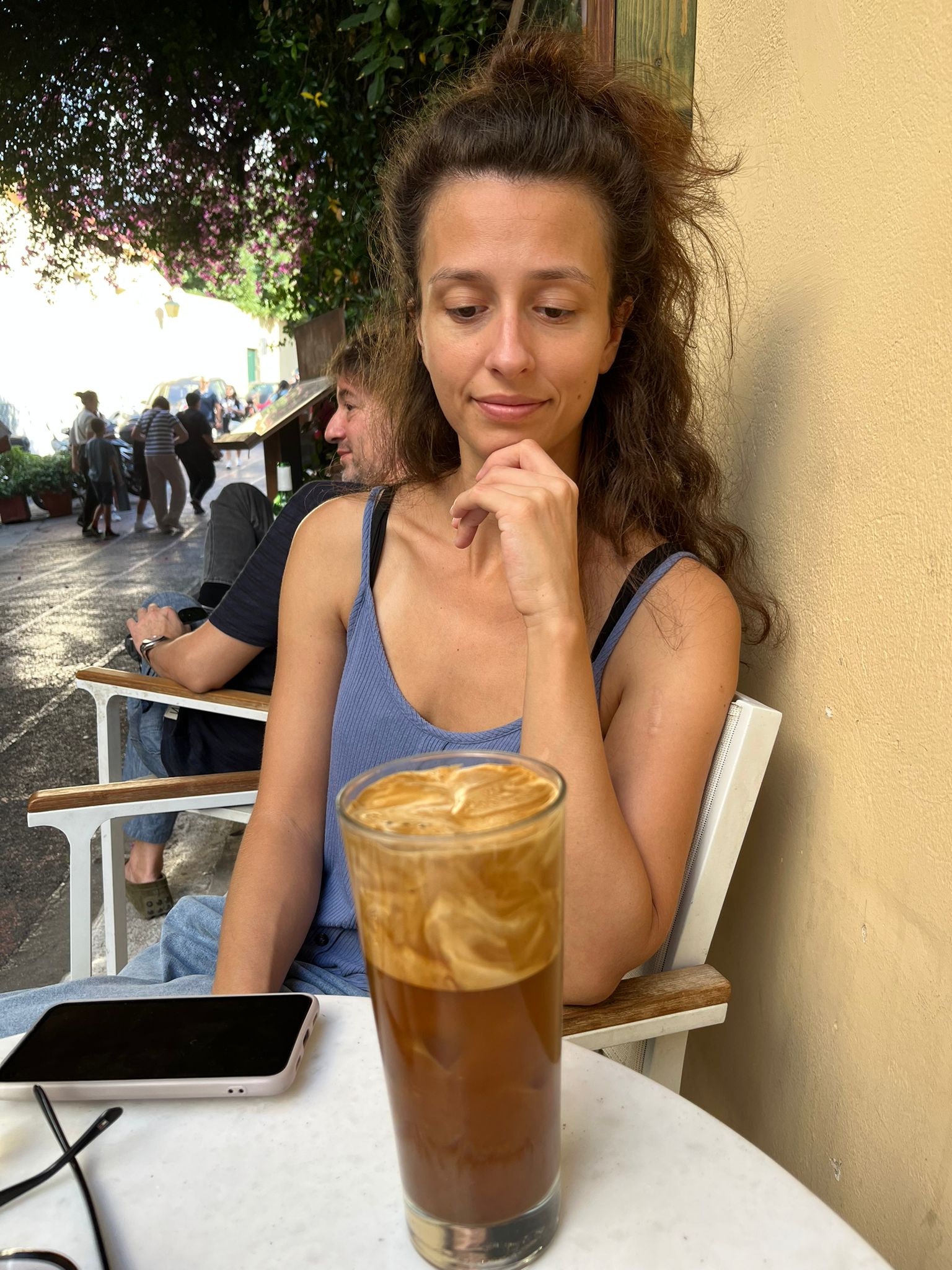 Dievča v modrom tričku sedí za stolom a pozerá sa na veľký pohár plný kávy. Na stole je aj mobil.