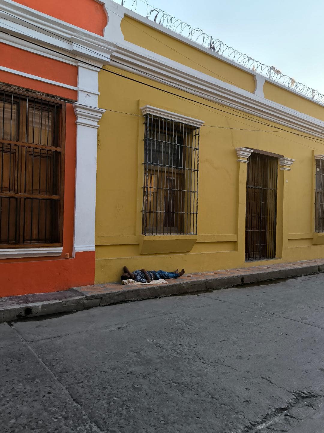Chlap leží na chodníku a spí. Za ním je žltý a oranžový dom.