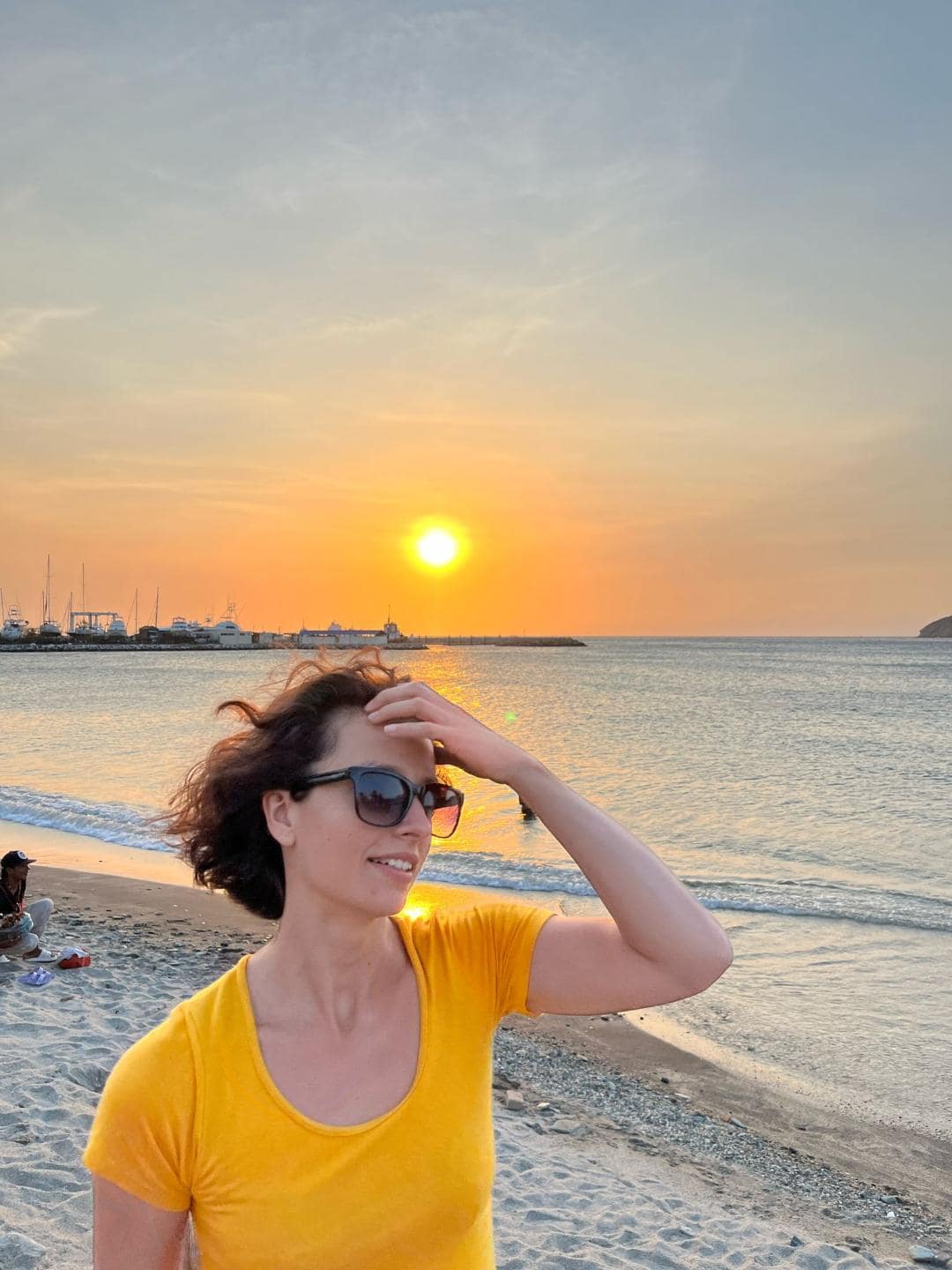 Dievča v oranžovom tričku stojí na pláži pri západe slnka a rukou sa hrabe vo vlasoch.