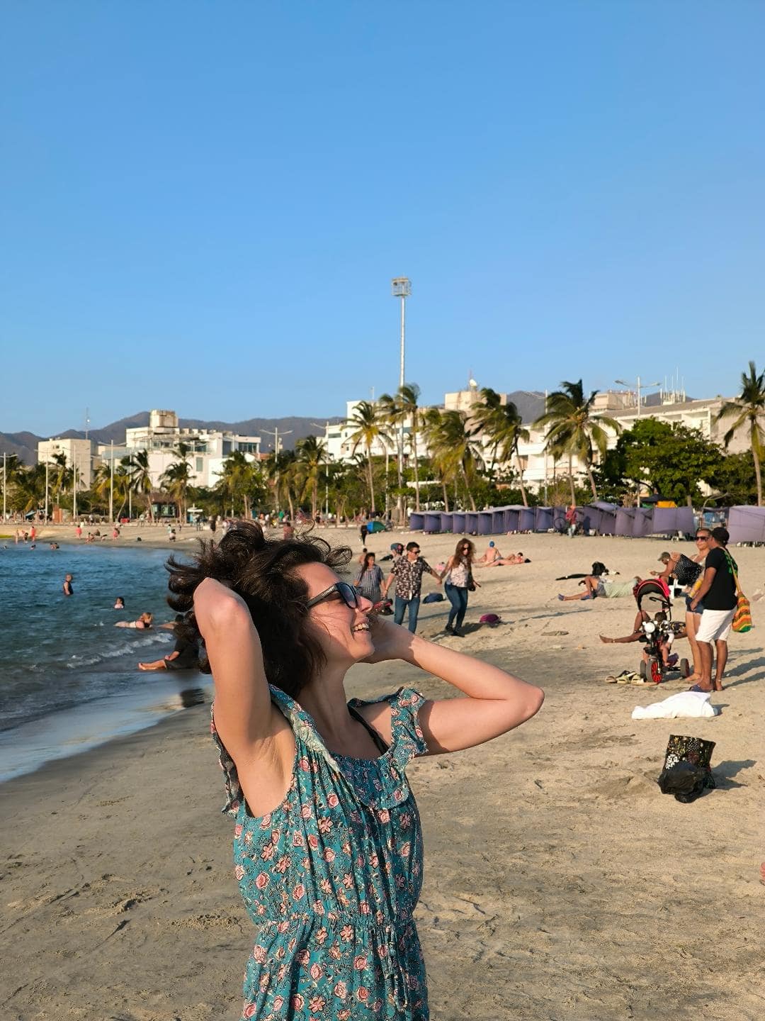 Dievča na pláži sa hrá s vlasmi. Za ňou sú turisti na pláži a vysoké palmy. Fúka silný vietor.