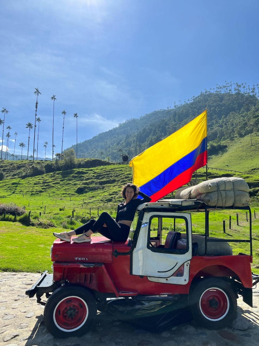 Dievča sedí na kapote červeného jeepu. Za ňou veje kolumbijská vlajka.