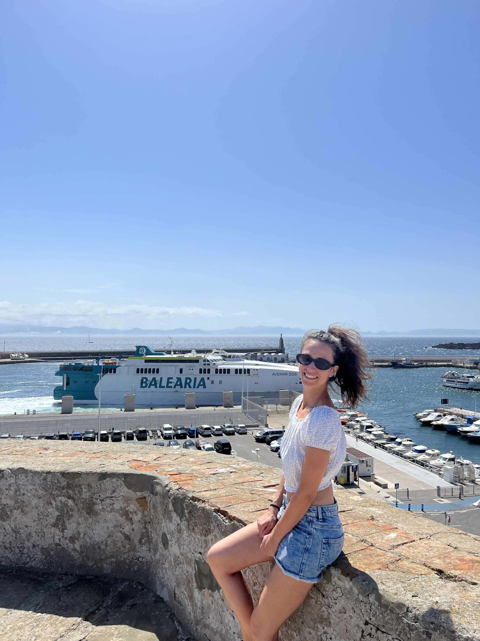 Dievča v bielej blúzke sedí na historických hradbách. Za ňou je more, na ktorom sa plaví veľká výletná loď.