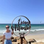 Dievča stojí pri kruhovej tyči pri mori. Za ňou lietajú kitesurferi.