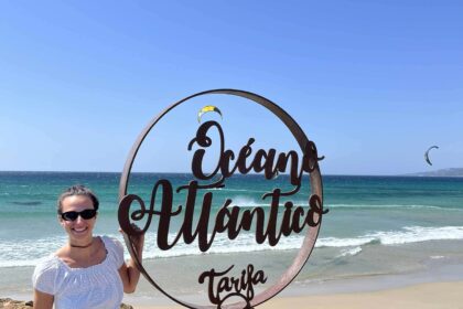 Dievča stojí pri kruhovej tyči pri mori. Za ňou lietajú kitesurferi.