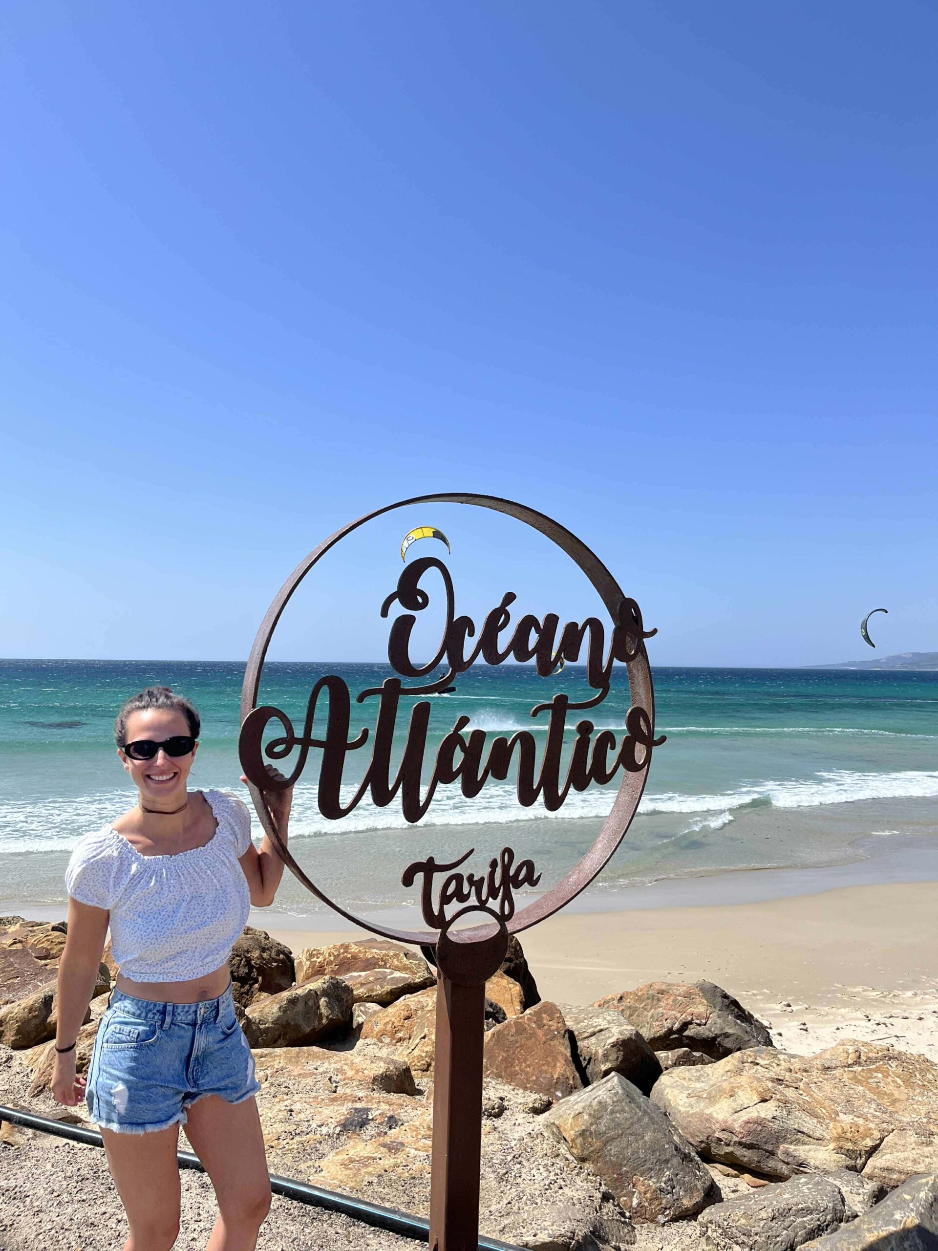 Dievča stojí pri kruhovej tyči pri mori. Za ňou lietajú kitesurferi.