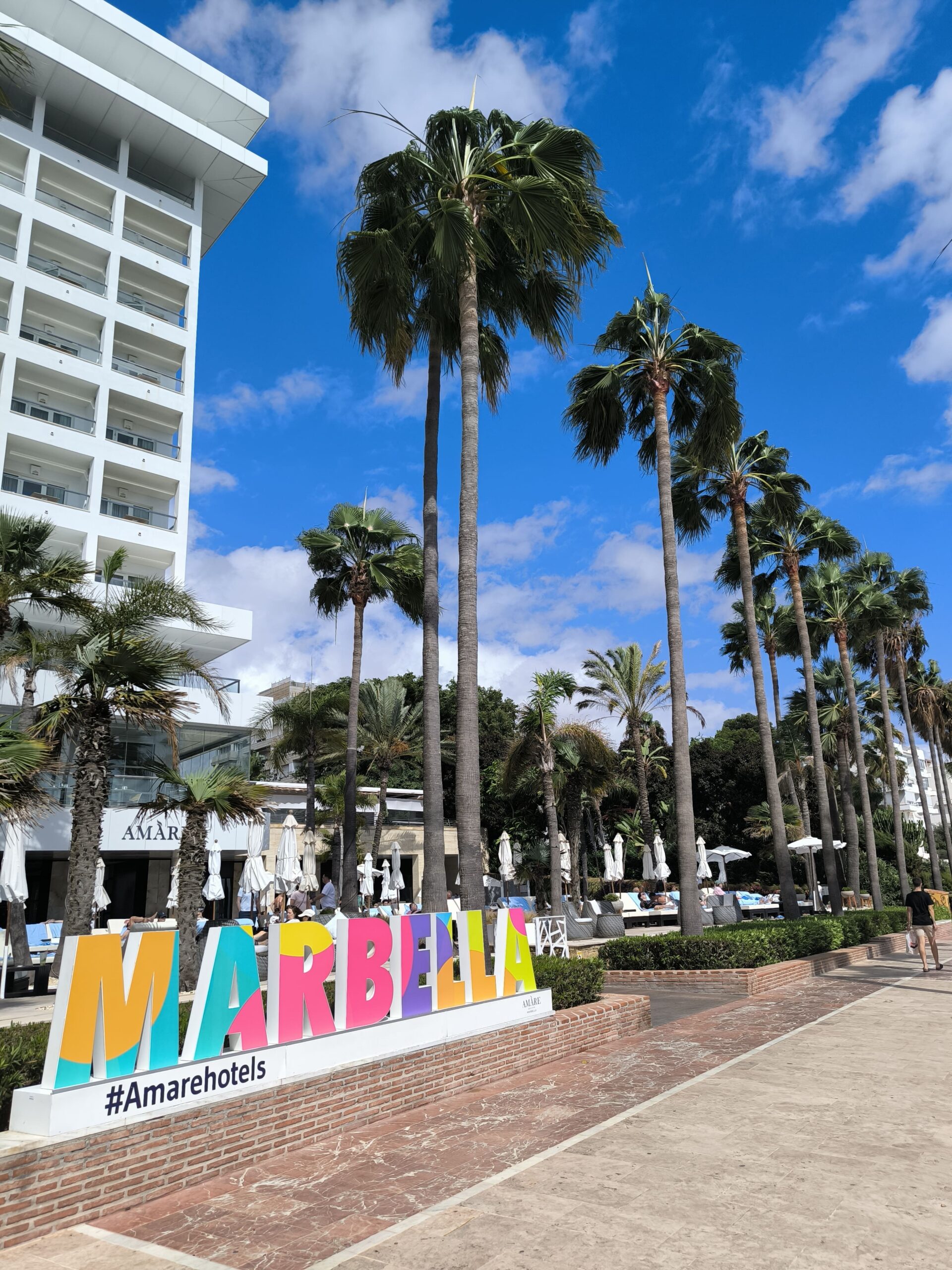 Popri promenáde stoja vysoké palmy a je tam veľký farebný nápis Marbella.