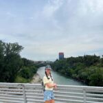 Dievča v žltom tričku a šiltovke stojí na moste. Pod ňou tečie rieka a v pozadí sú lesy.